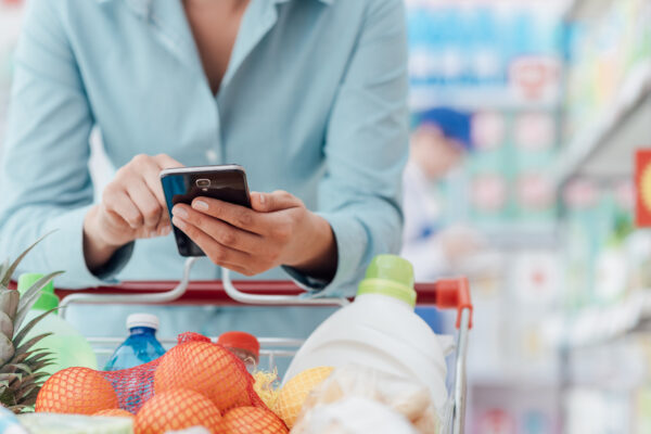 Woman doing grocery shopping at the supermarket, she is leaning on the shopping cart and connecting with her phone, apps and retail concept