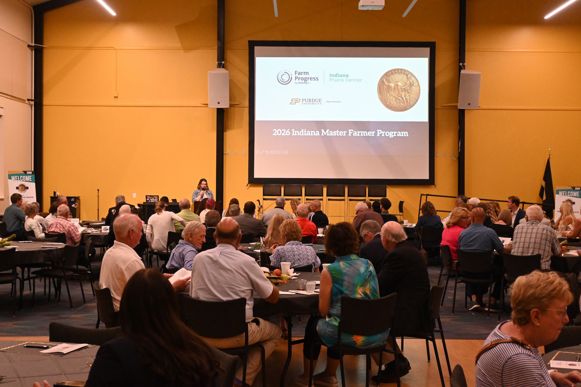 Conference room with seated attendees, speaker at podium and large screen reading "2026 Indiana Master Farmer Program."