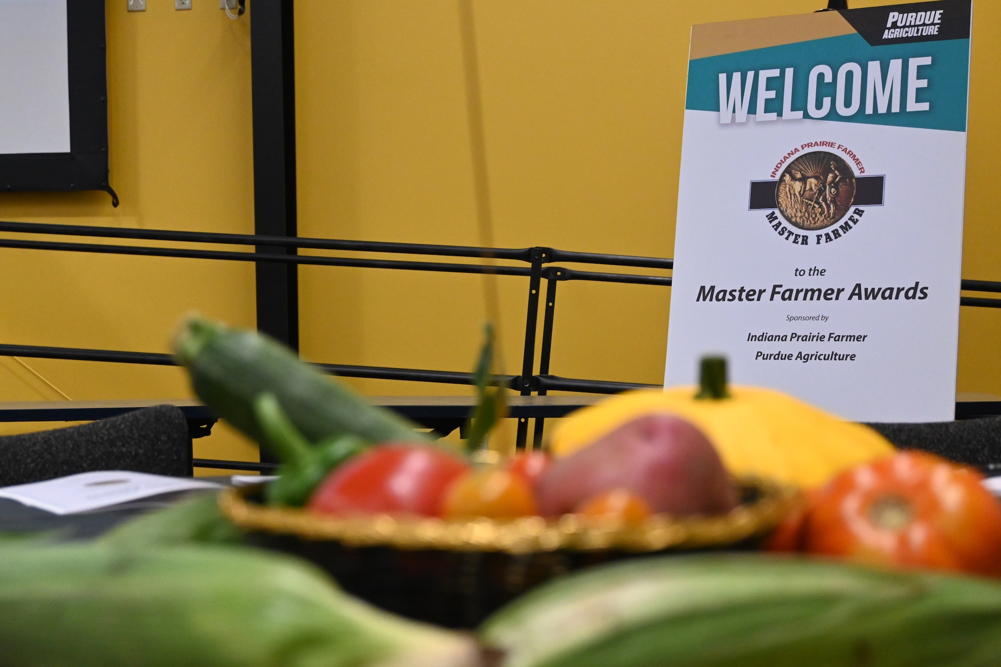 Welcome sign for Master Farmer Awards (Purdue Agriculture) with blurred vegetables in foreground.