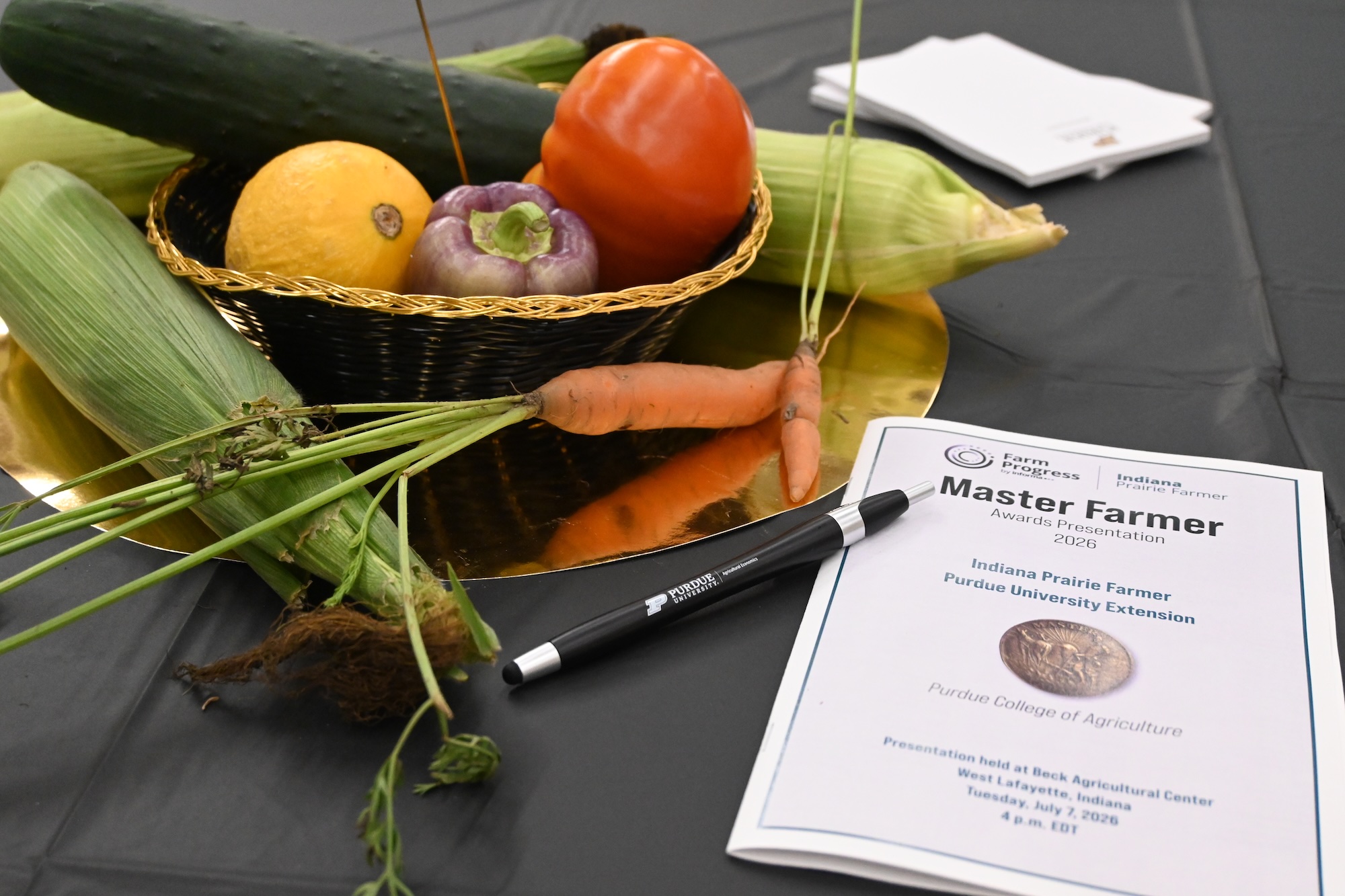 Basket of vegetables with corn and carrots beside a "Master Farmer" awards booklet and a Purdue pen.