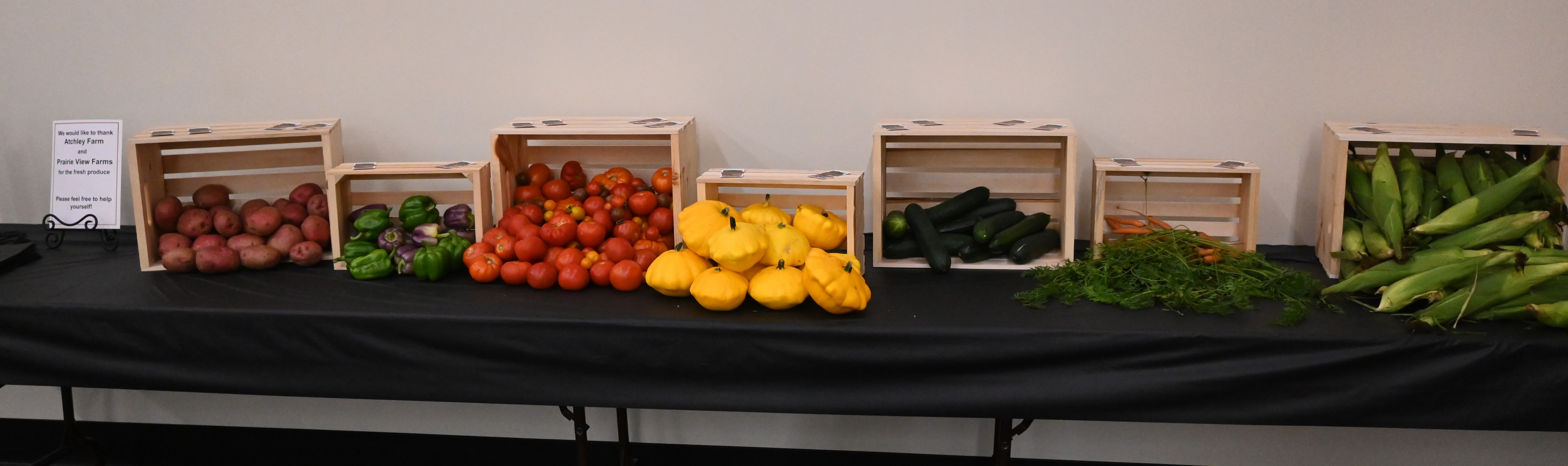 Wooden crates on a table holding potatoes, bell peppers, tomatoes, yellow pattypan, zucchini, carrots and corn.