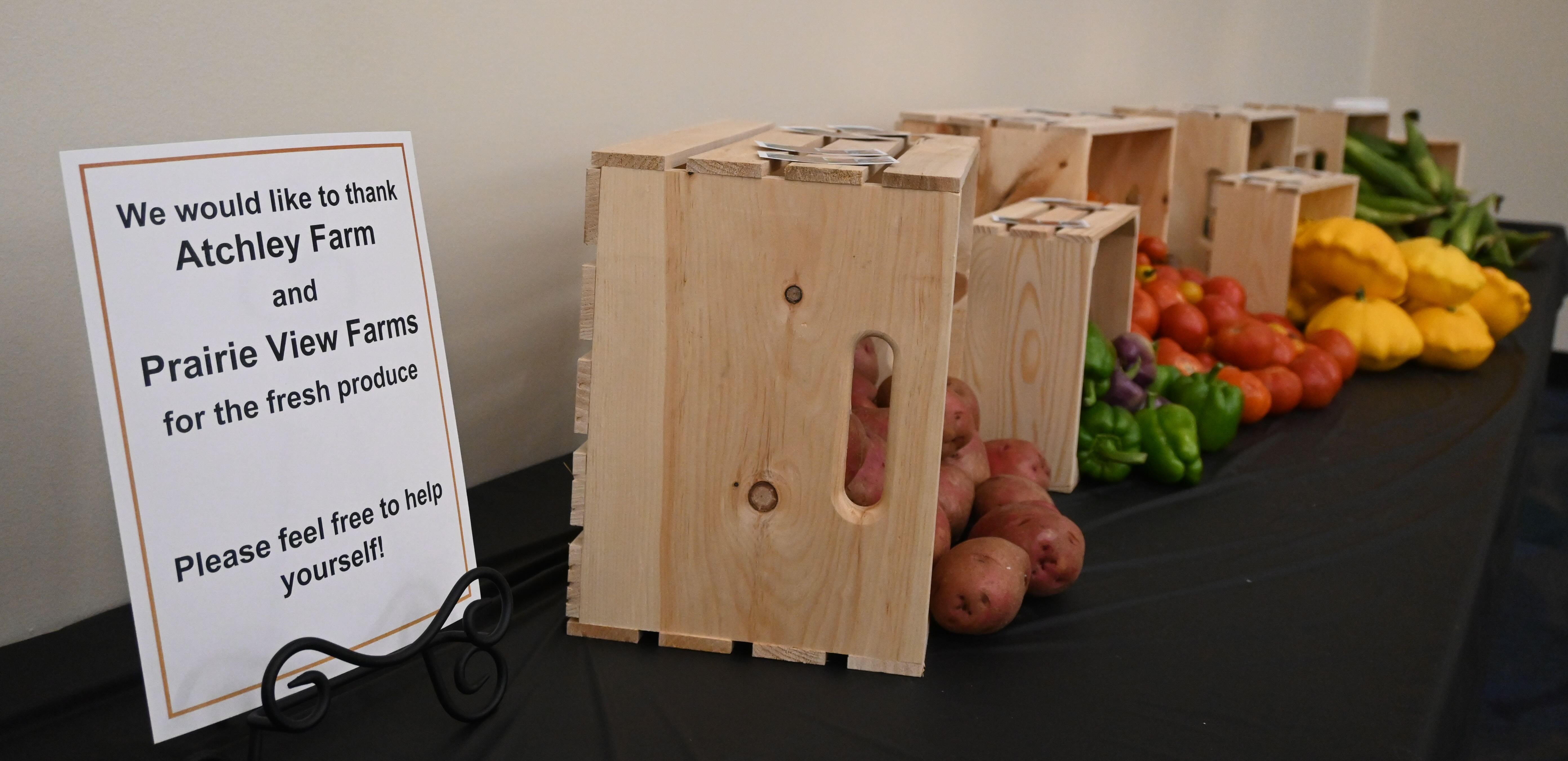 Sign thanking Atchley Farm and Prairie View Farms beside wooden crates of mixed produce on a table.