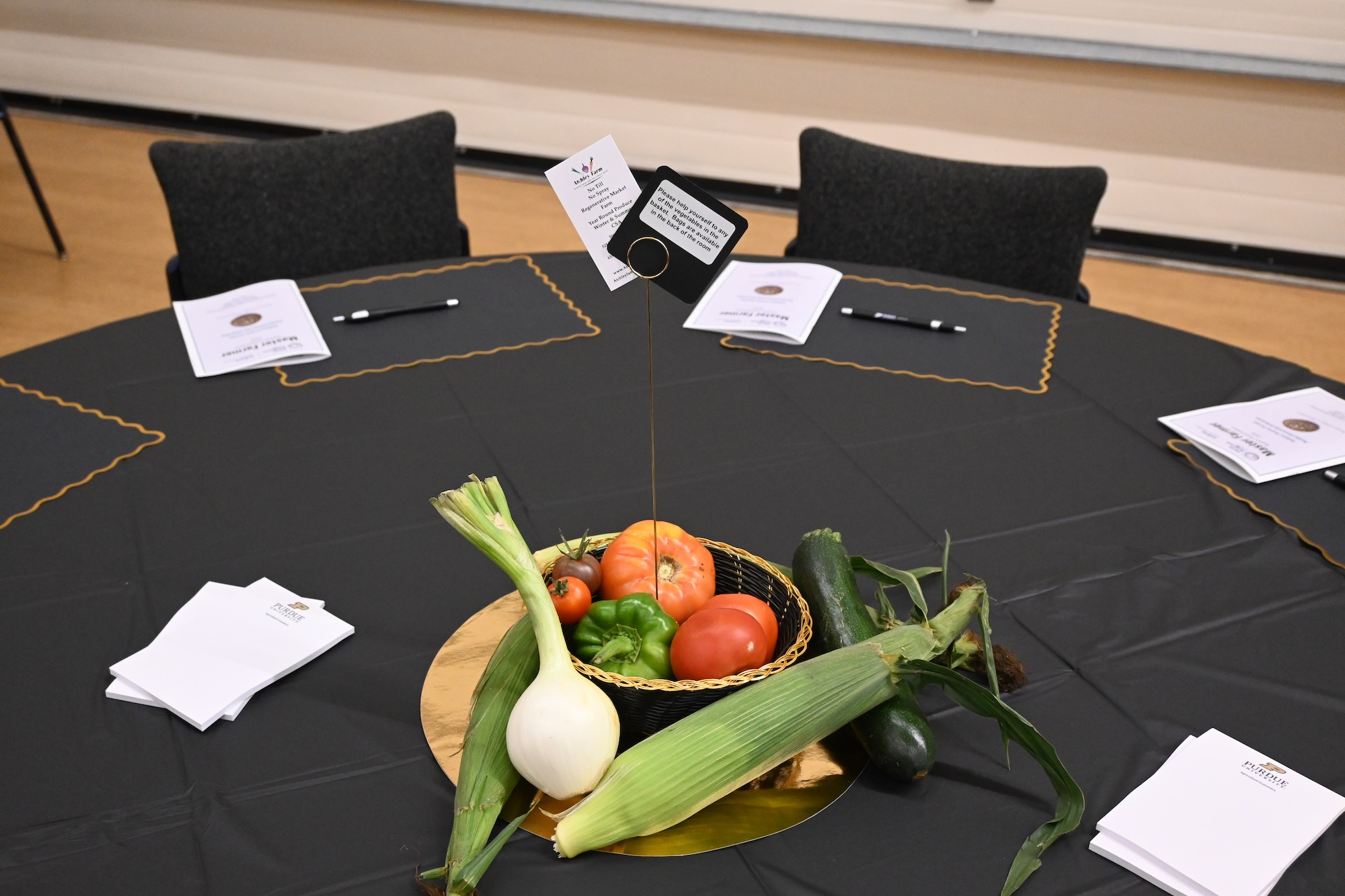 Black-draped round table with vegetable centerpiece (onion, corn, tomatoes, green pepper, zucchini) and booklets with pens.