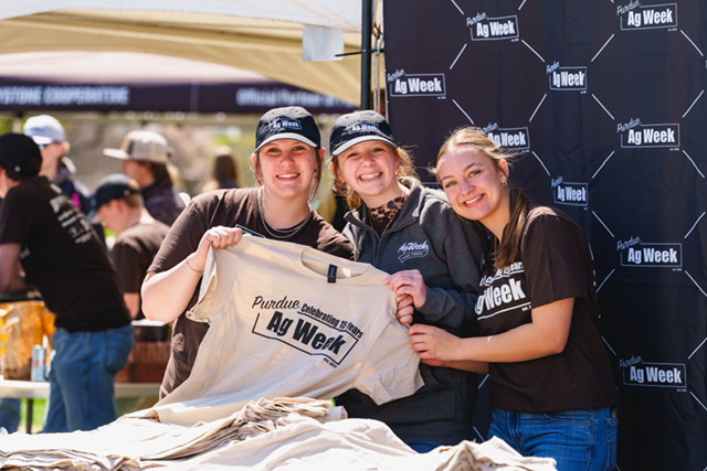 Anglemyer with friends in front of Ag Week banner holding and Ag Week shirt