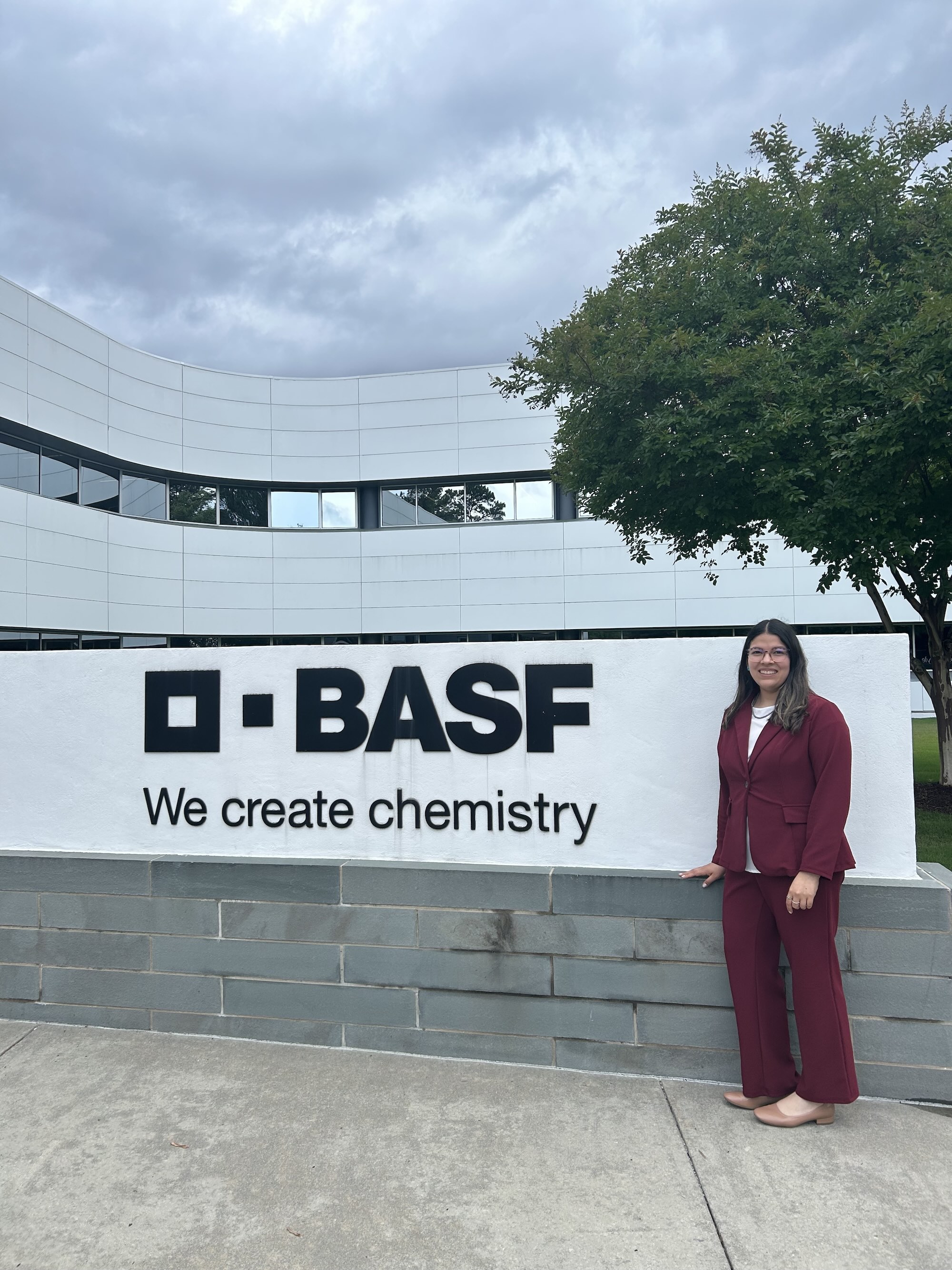 Berenize standing in a red suit in front of BASF sign that says "we create chemistry"