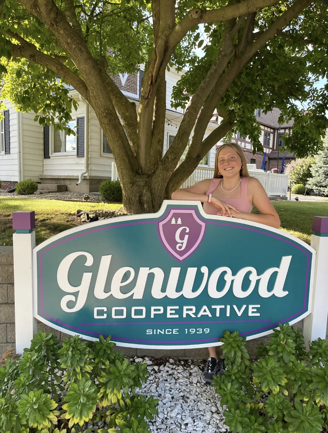Emily Recker standing behind the Glenwood Cooperative sign in front of the cooperative house