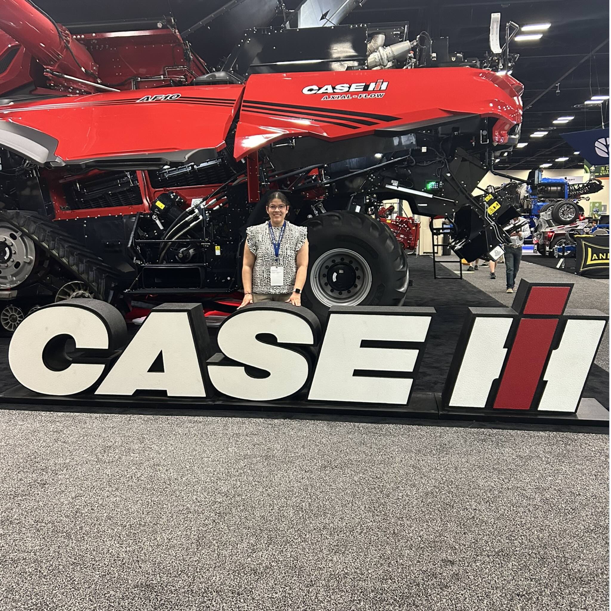 Berenize standing behind a large white CASE IH sign in front of a red Case IH combine harvester at an indoor expo
