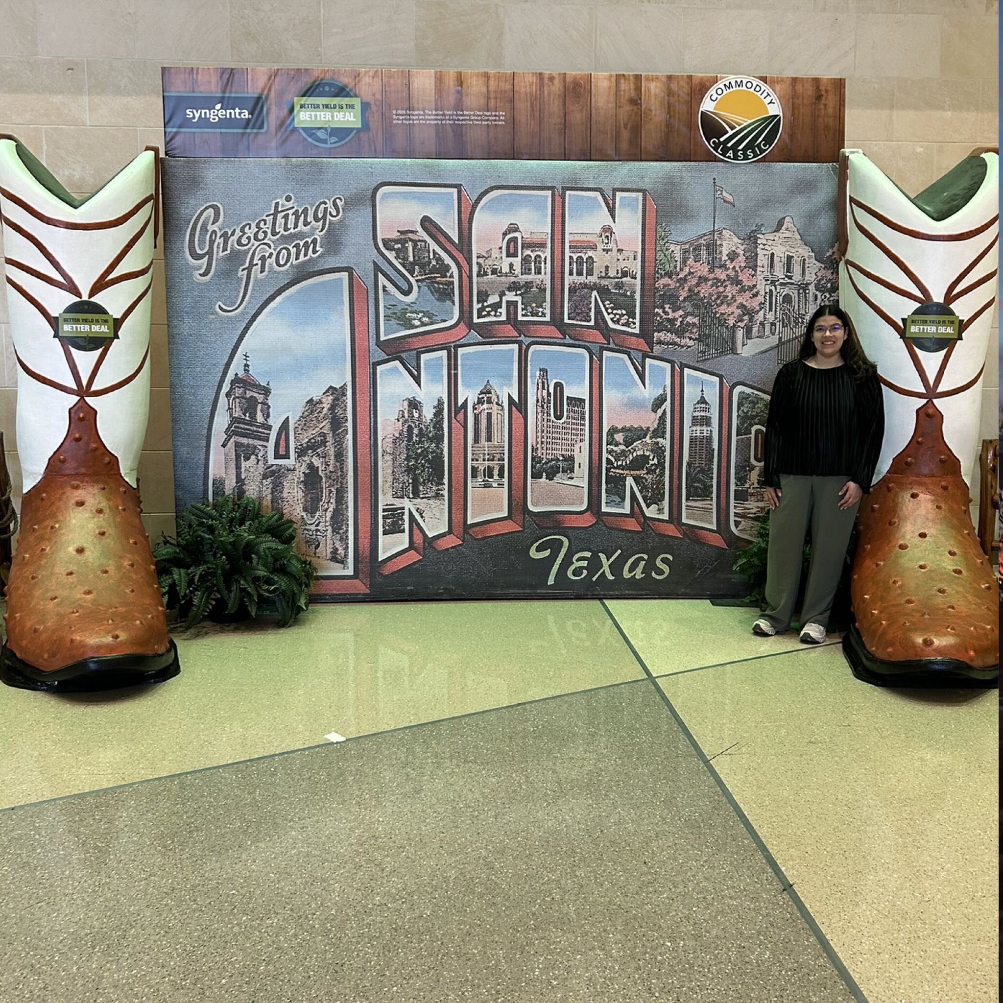 Berenize between oversized cowboy boots in front of a postcard backdrop reading Greetings from San Antonio
