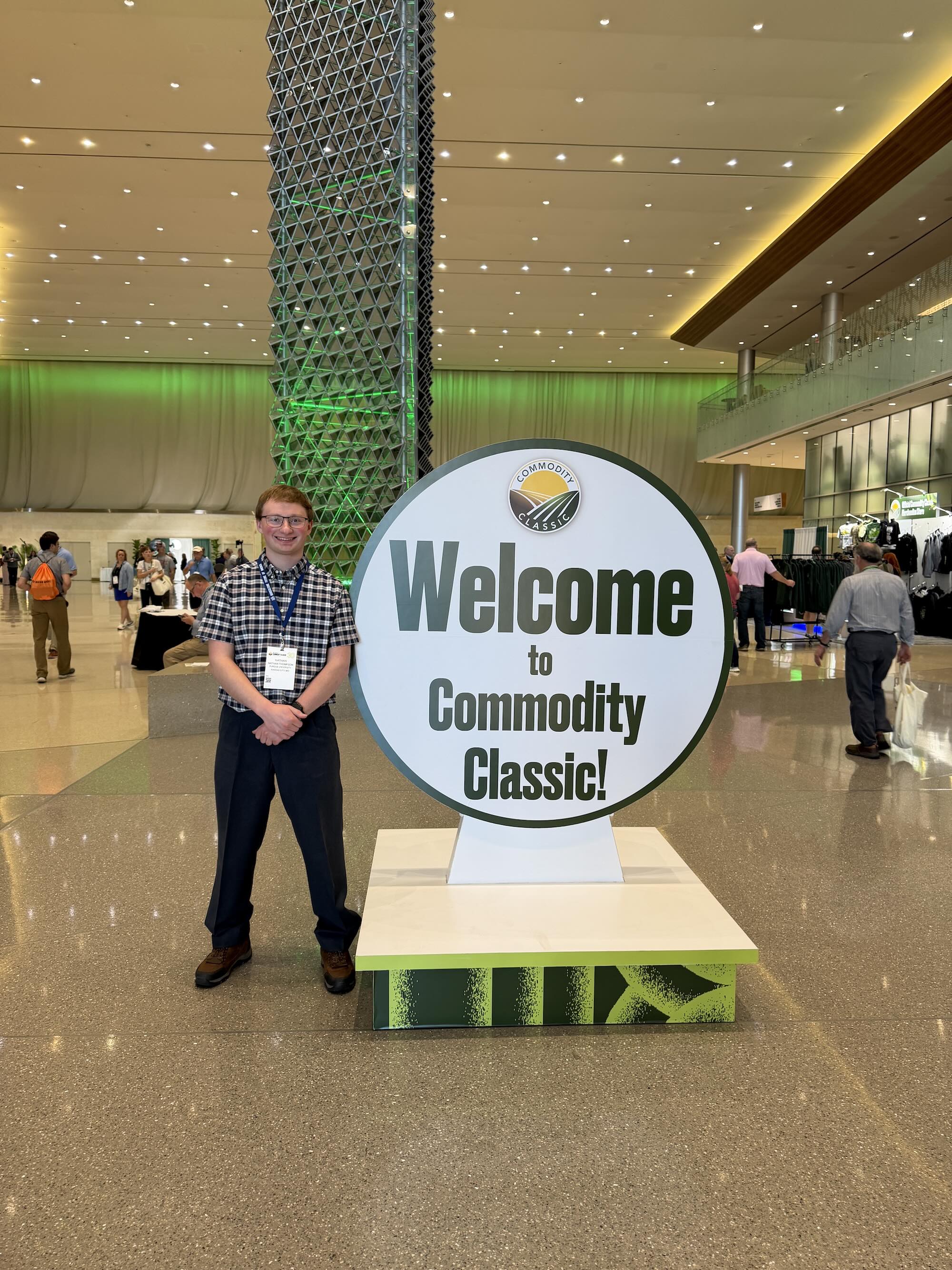 Nathan standing with conference badge beside a large round welcome sign for Commodity Classic in a convention hall