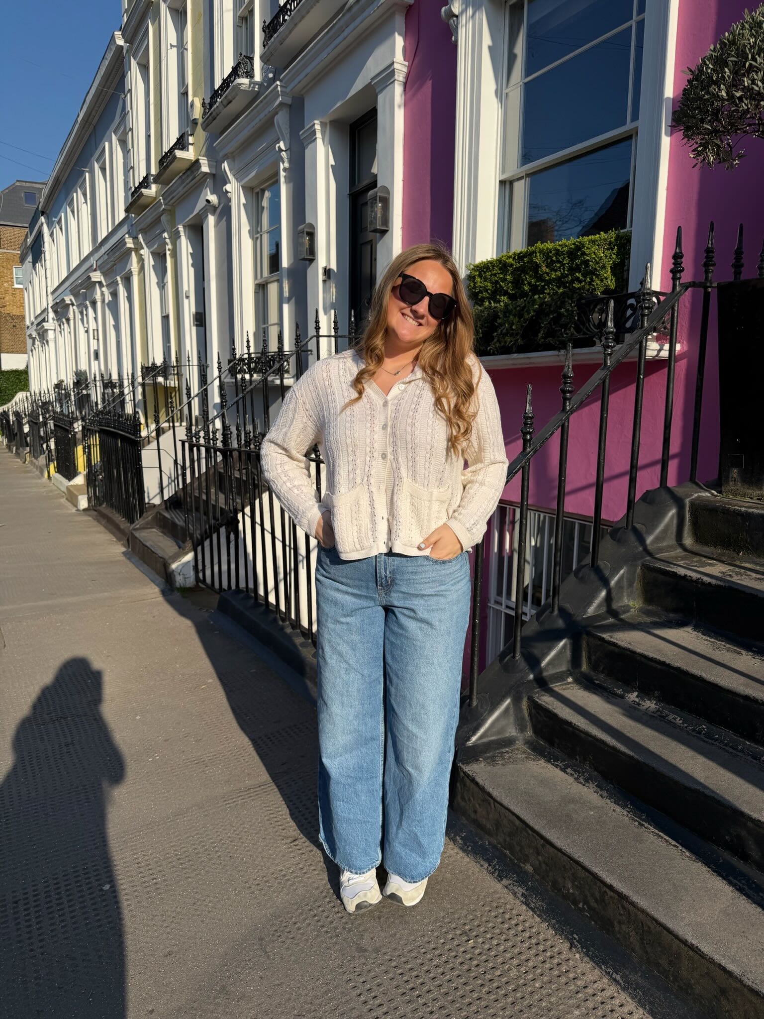 Klinkhamer smiling in sunglasses and white knit cardigan, standing by a pink townhouse with iron railings.