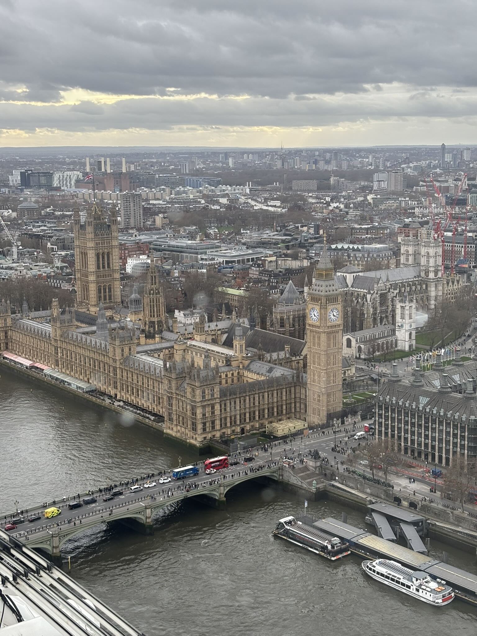 Houses of Parliament and Big Ben beside the Thames with Westminster Bridge, traffic and boats under a cloudy sky.