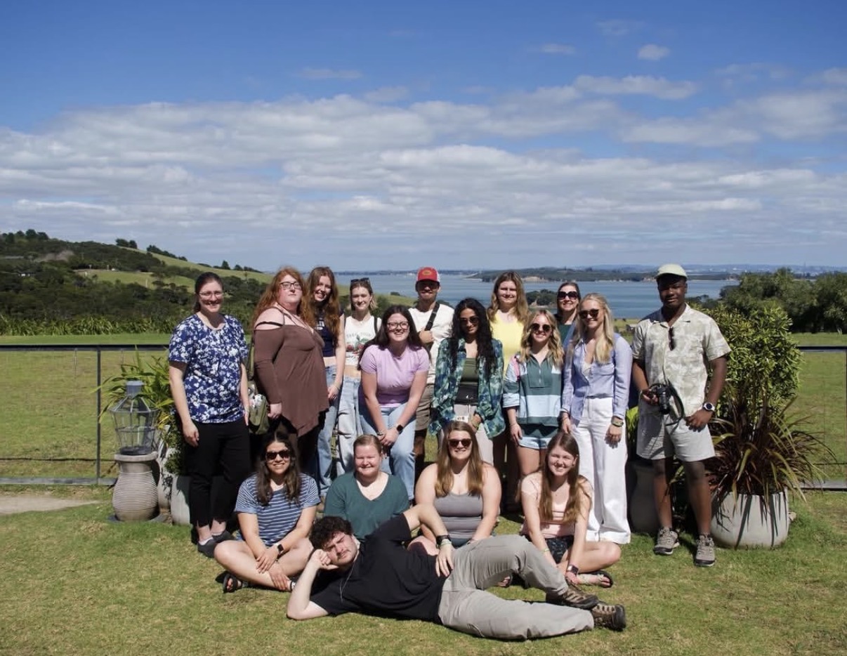 New Zealand study-abroad group posing on a grassy hill, with a coastal bay and rolling hills in the background.