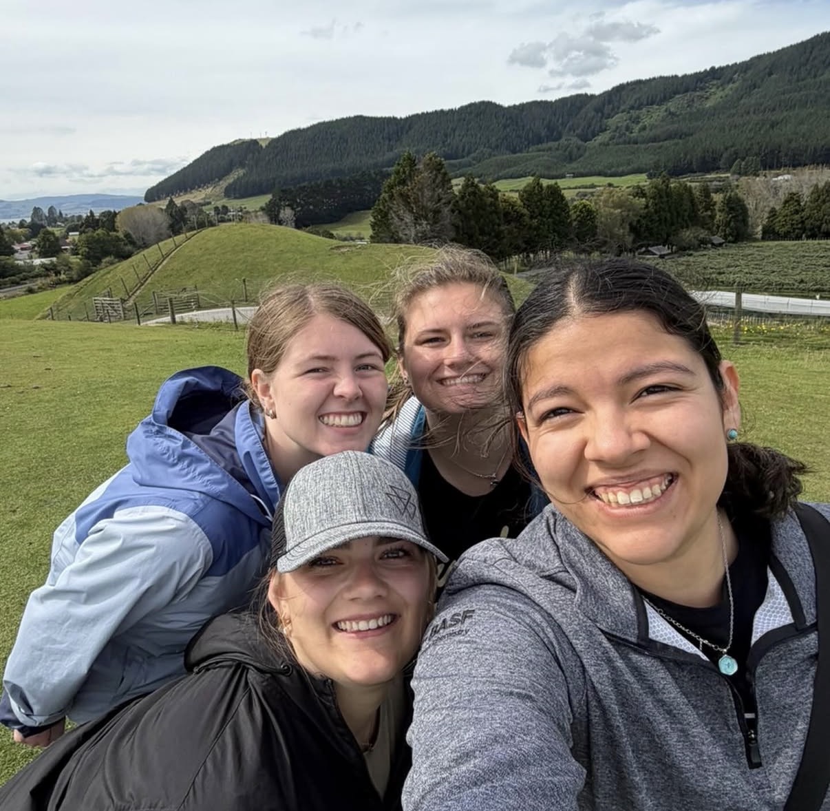 Four friends taking a selfie on a grassy hill with rolling green hills and trees in the background.