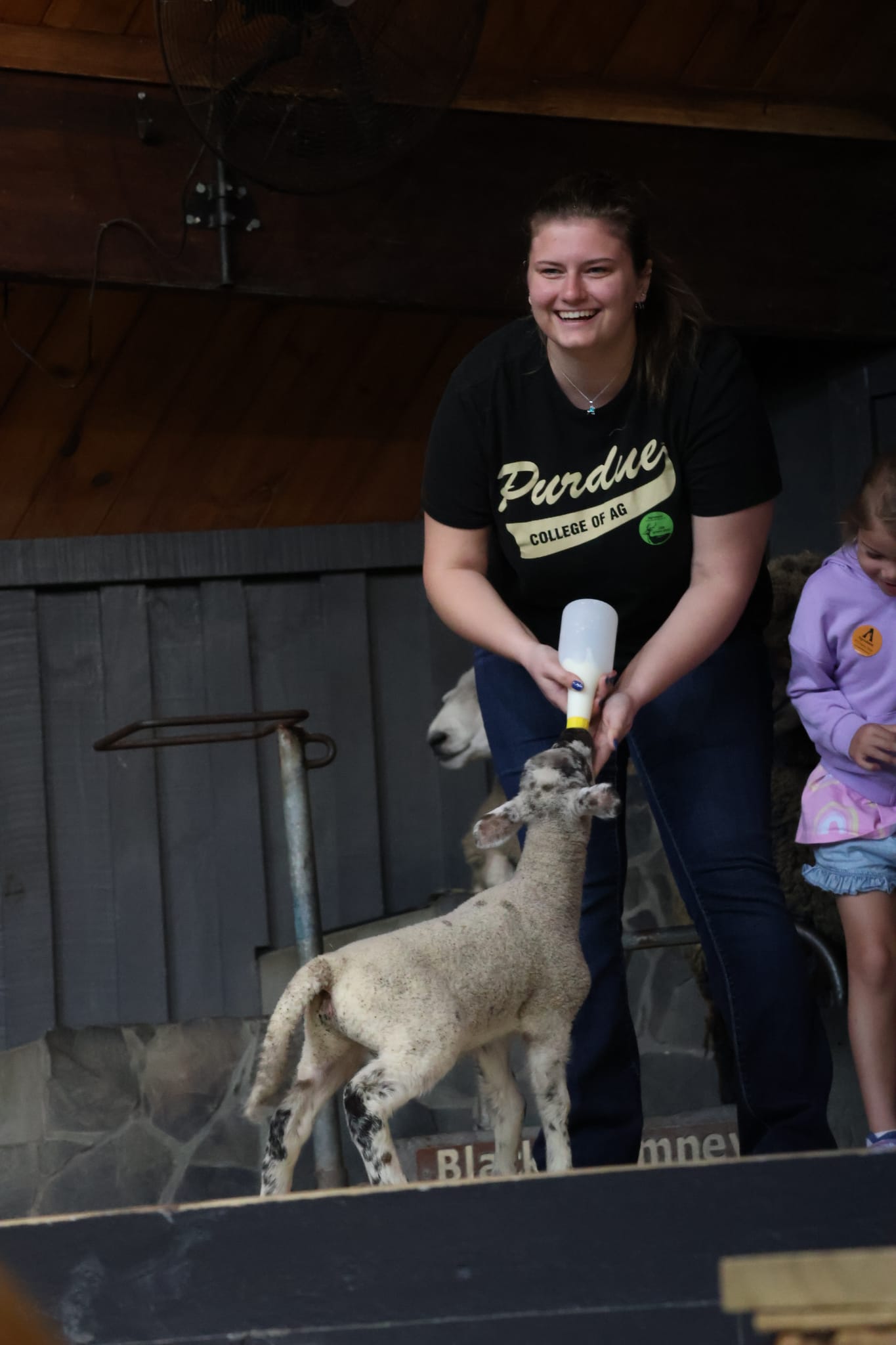 Woman wearing a Purdue College of Ag T‑shirt smiling as she bottle-feeds a lamb inside a barn
