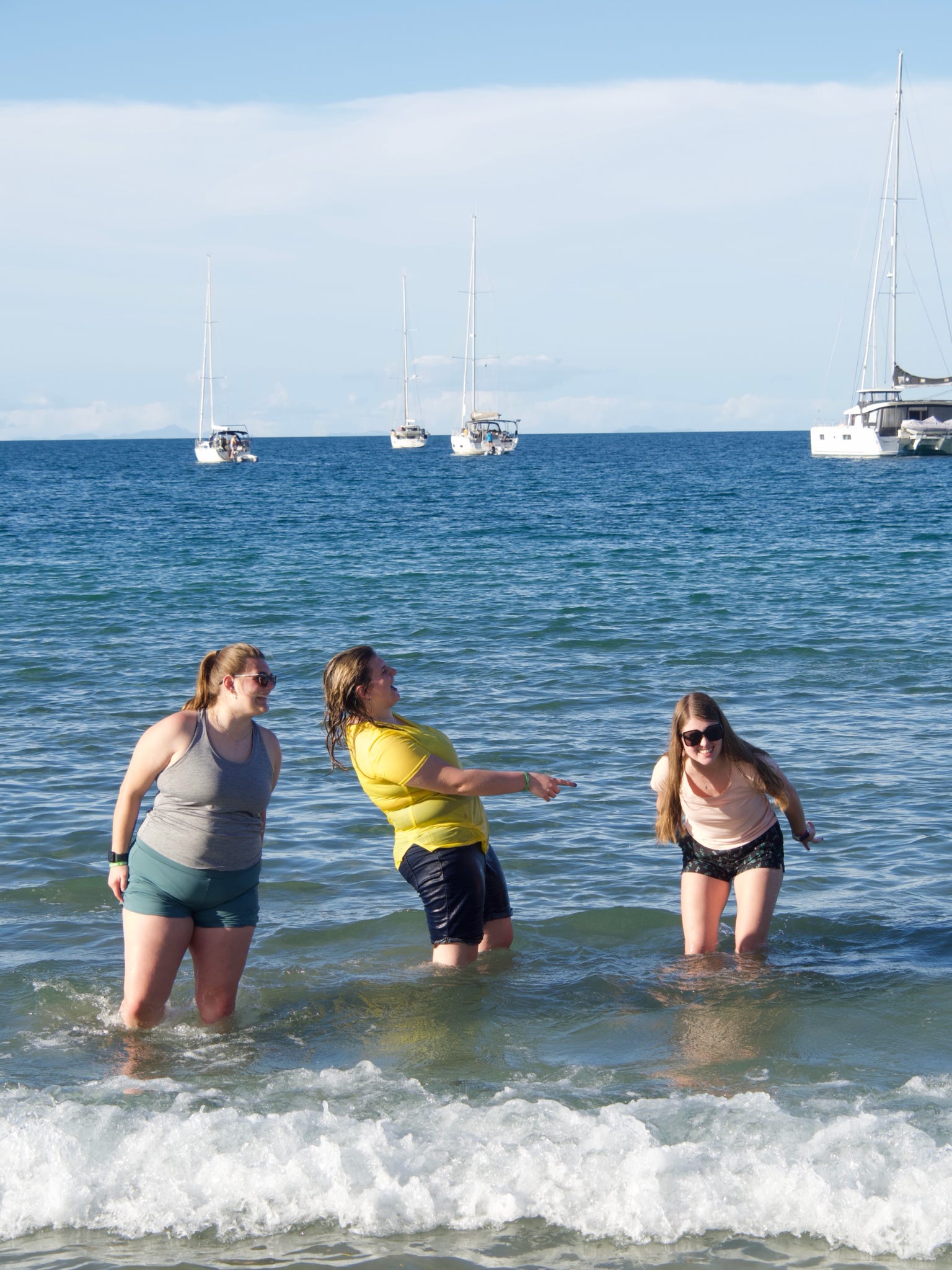 Three young women laughing and wading in shallow ocean water with sailboats anchored on the horizon.