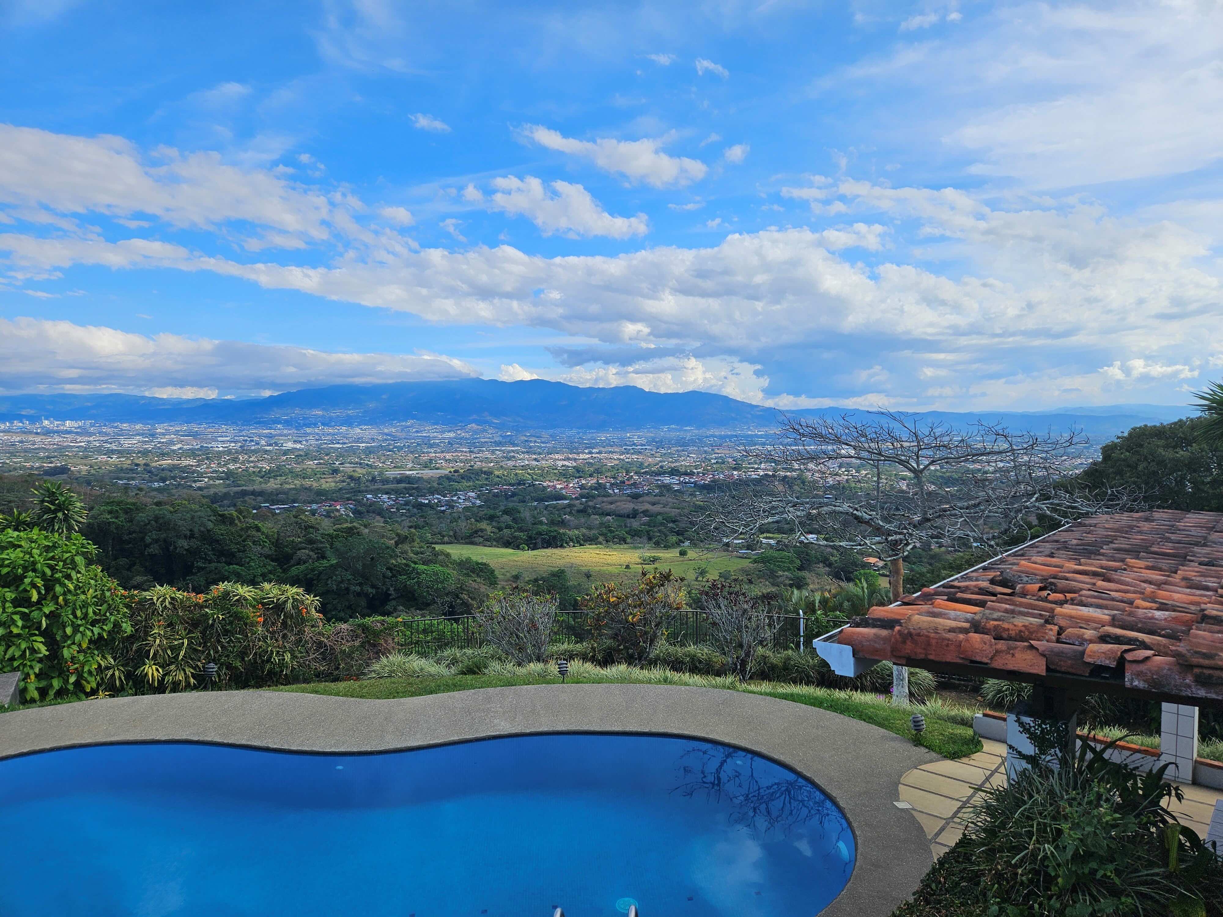Curved backyard pool overlooking green valley, distant city and blue mountains under a bright sky with scattered clouds.