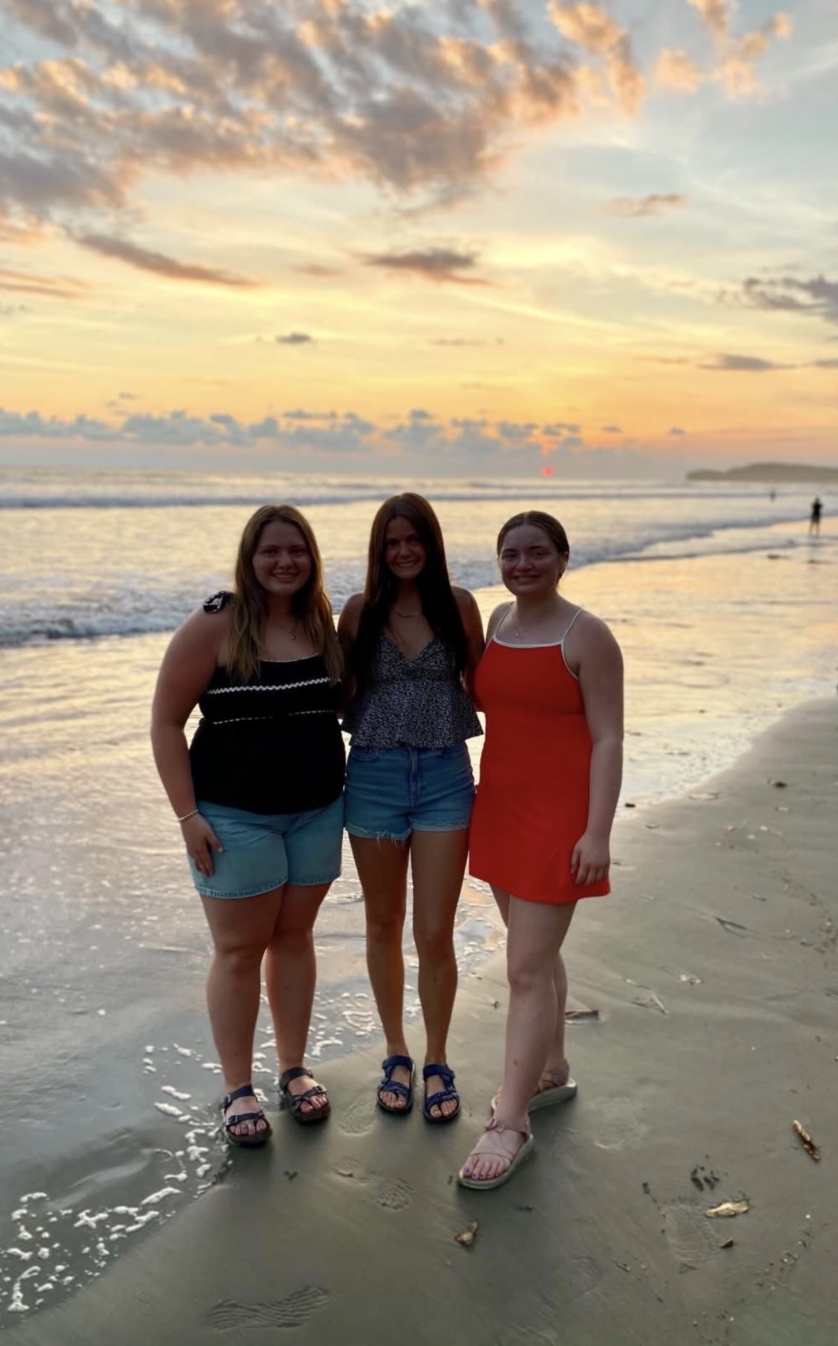 Three young women standing on a wet sandy beach at sunset, smiling with ocean waves and a colorful sky behind them.