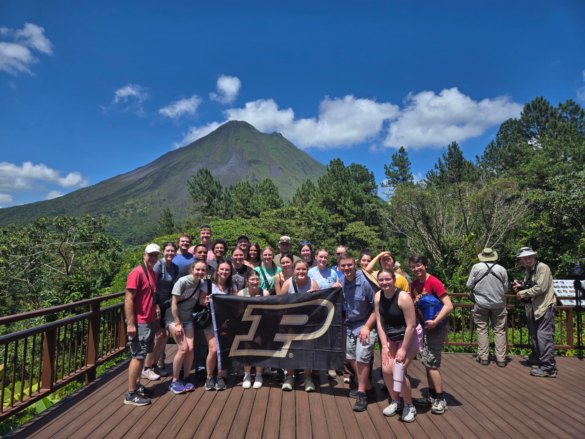 Group of people on a wooden viewing deck holding a Purdue banner with a mountain and blue sky in the background.