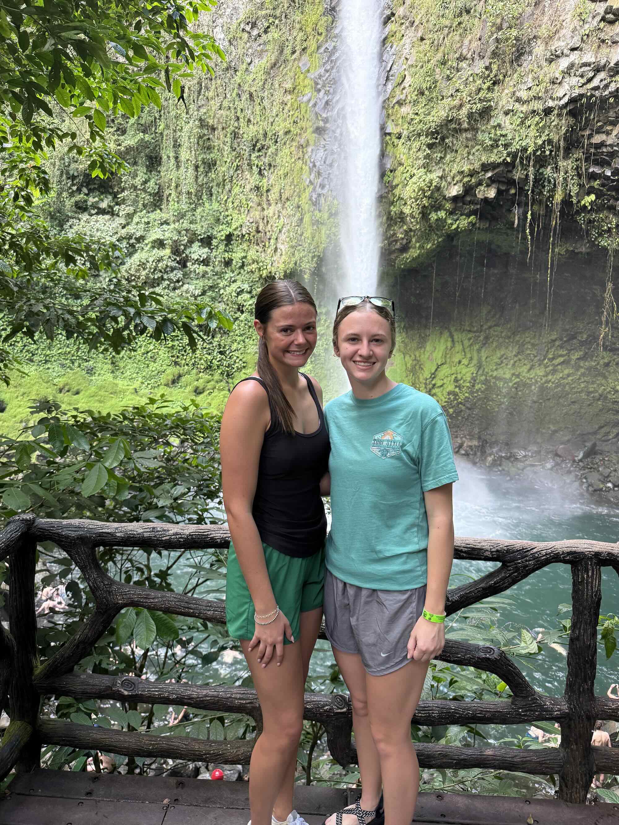 Kiersten and a friend are standing at a wooden railing with a tall waterfall and lush green cliff behind them.