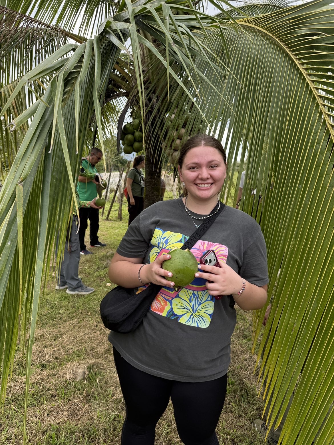 Lillian is under palm fronds, holding a green coconut, with people and a coconut tree in the background.