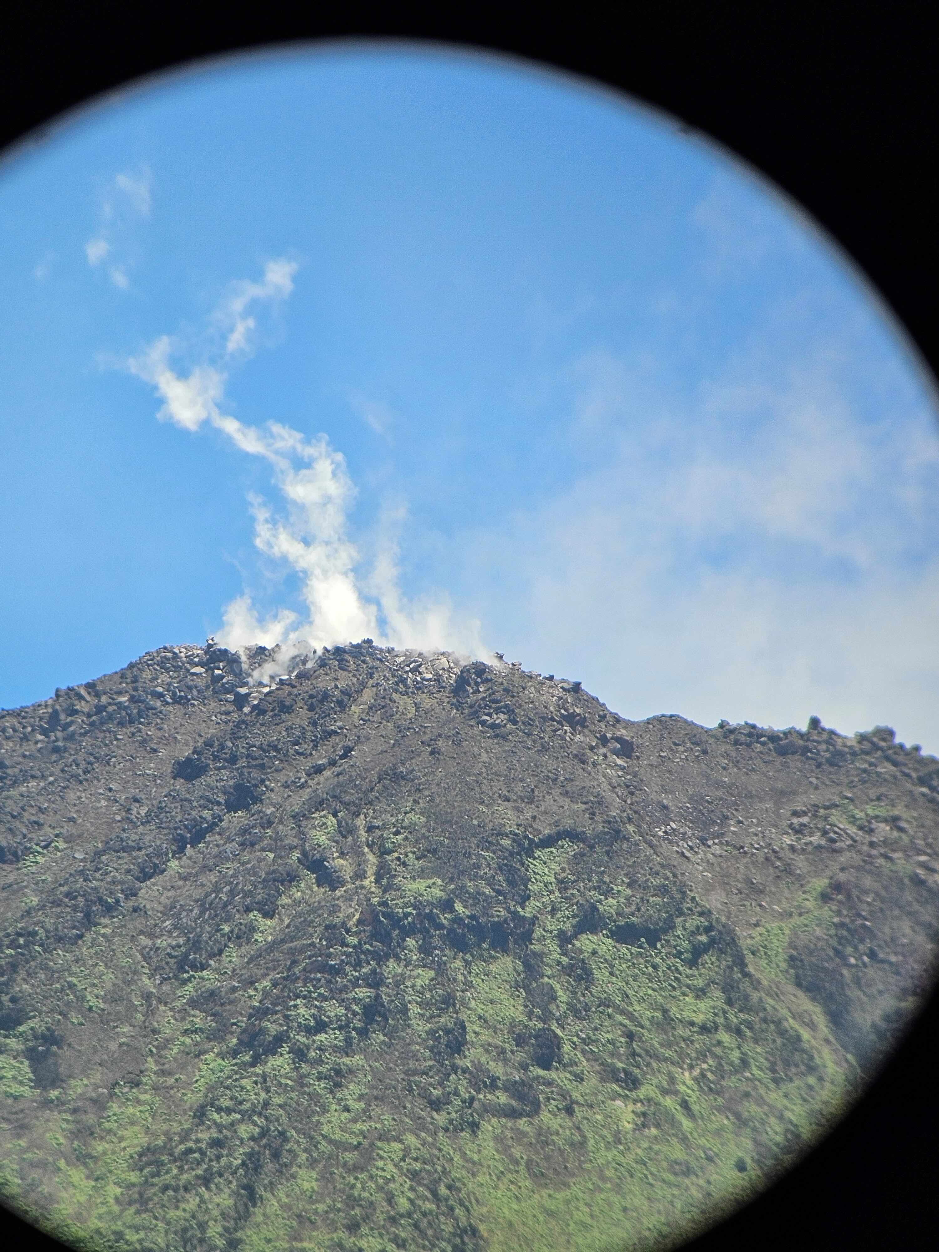 Steep volcano with thin white steam rising from the summit under a clear blue sky, seen through a circular lens.