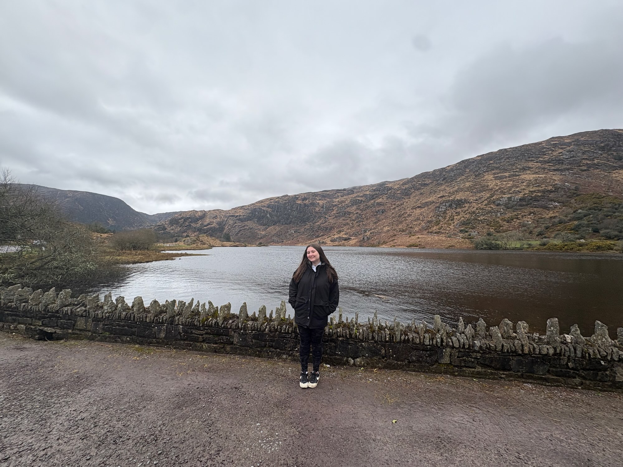 Emma, in a black coat, stands by a stone wall beside a calm lake with brown hills and a cloudy sky.