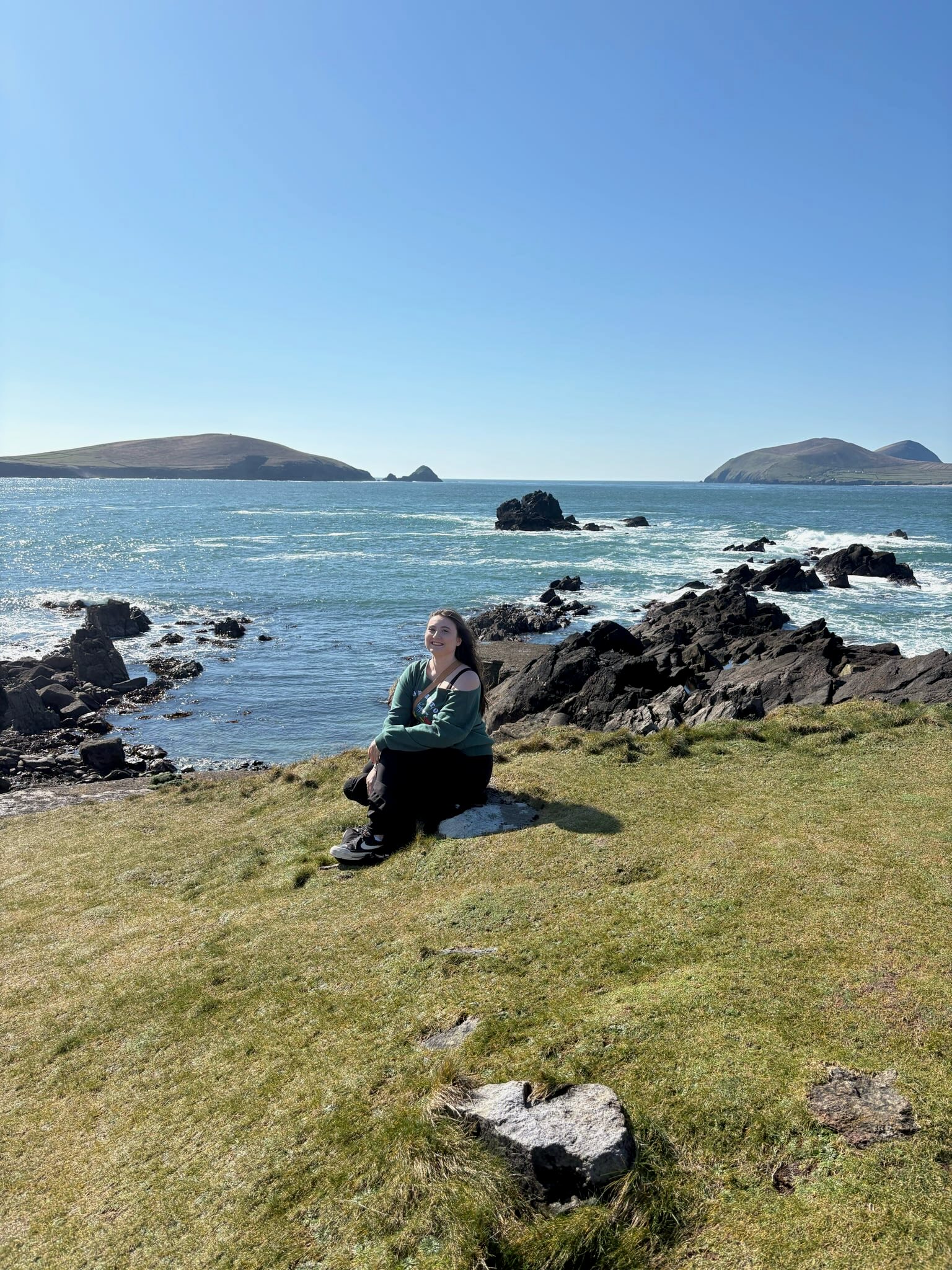 Emma is sitting on a grassy cliff by a rocky shoreline, smiling toward a calm blue sea with distant islands.