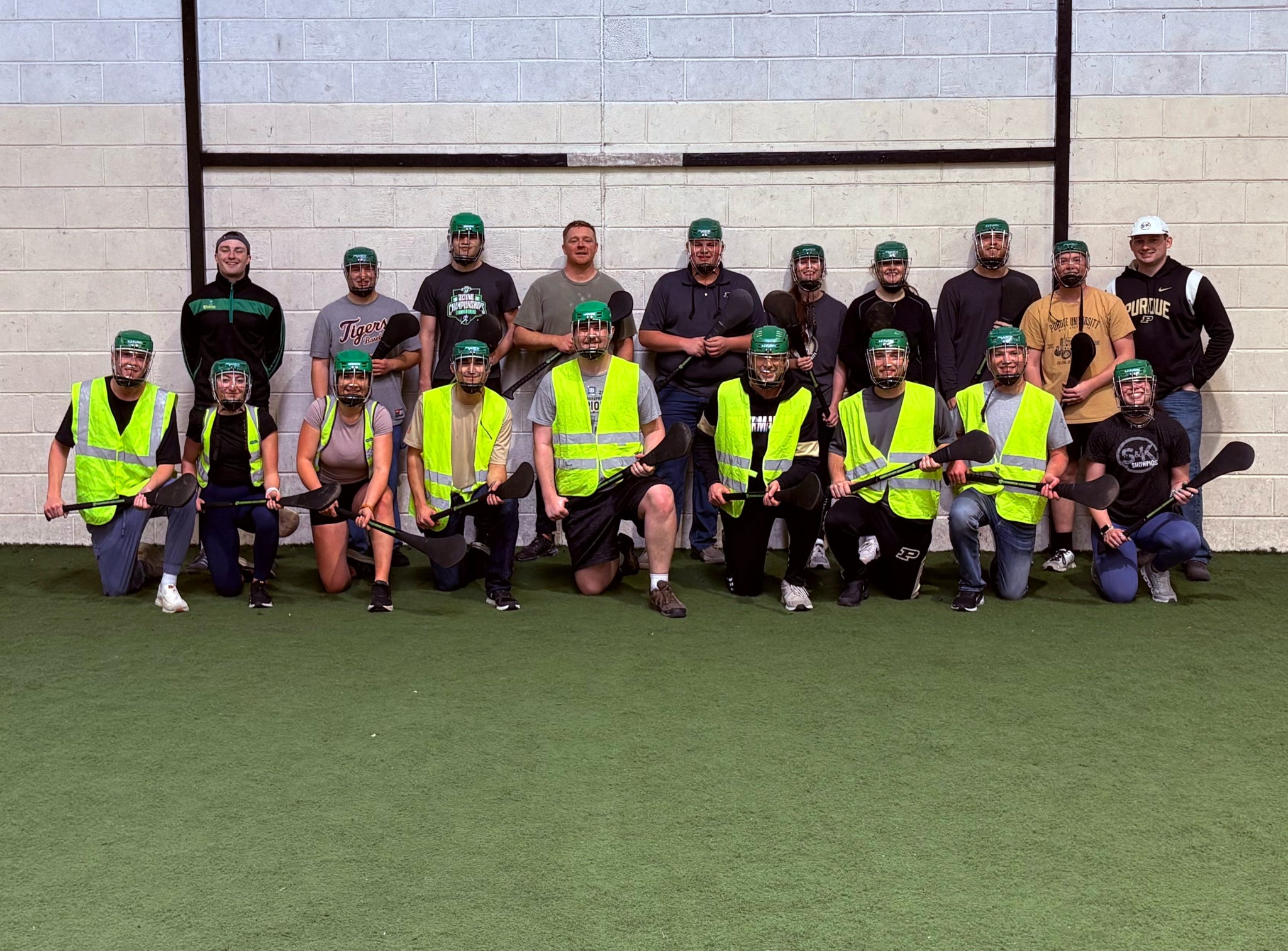 Students wearing green helmets and holding hurling sticks, some in neon safety vests, posed inside a gym.