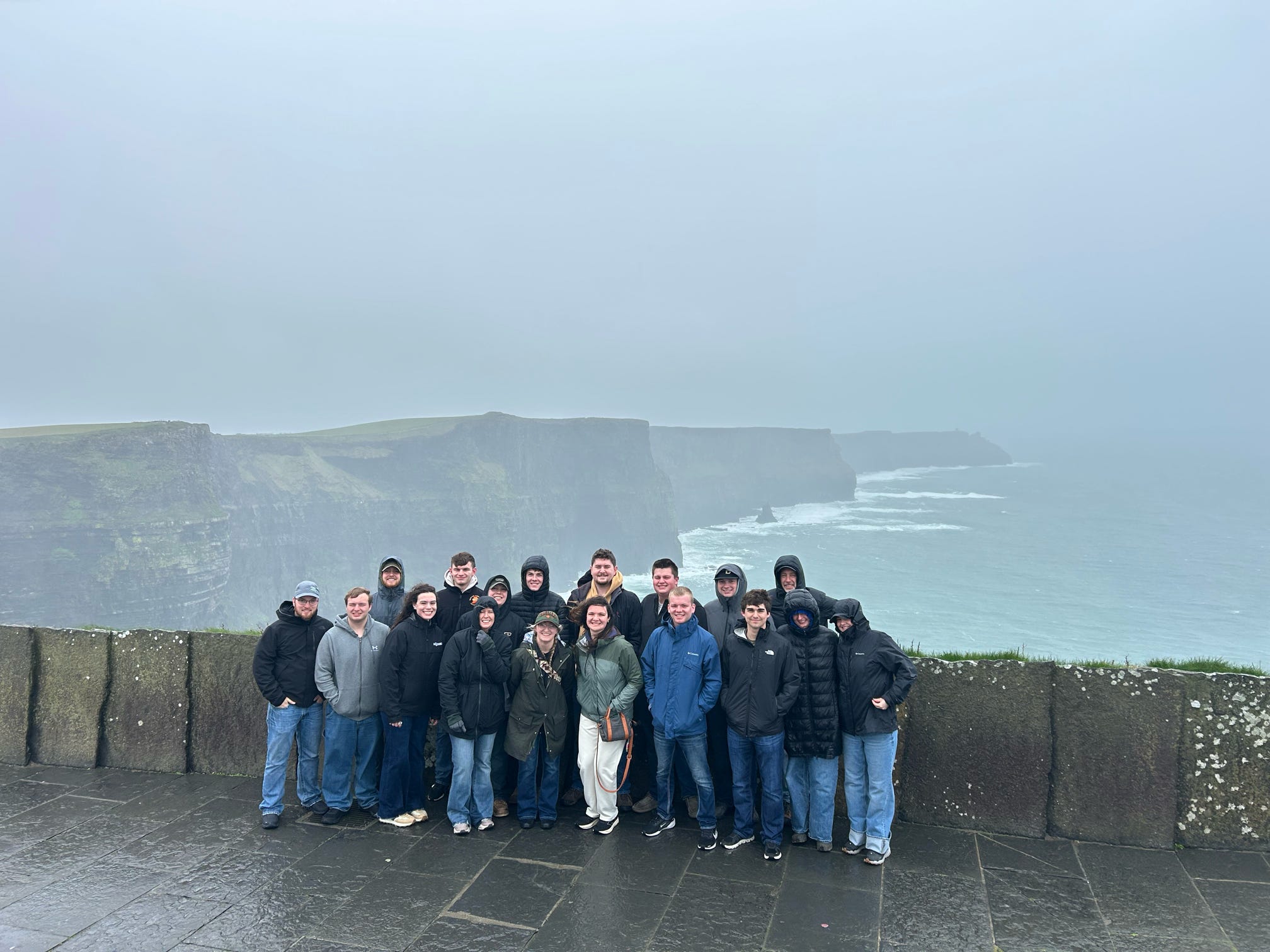 Ireland study-abroad group standing on a stone wall, with misty sea cliffs and waves behind them.