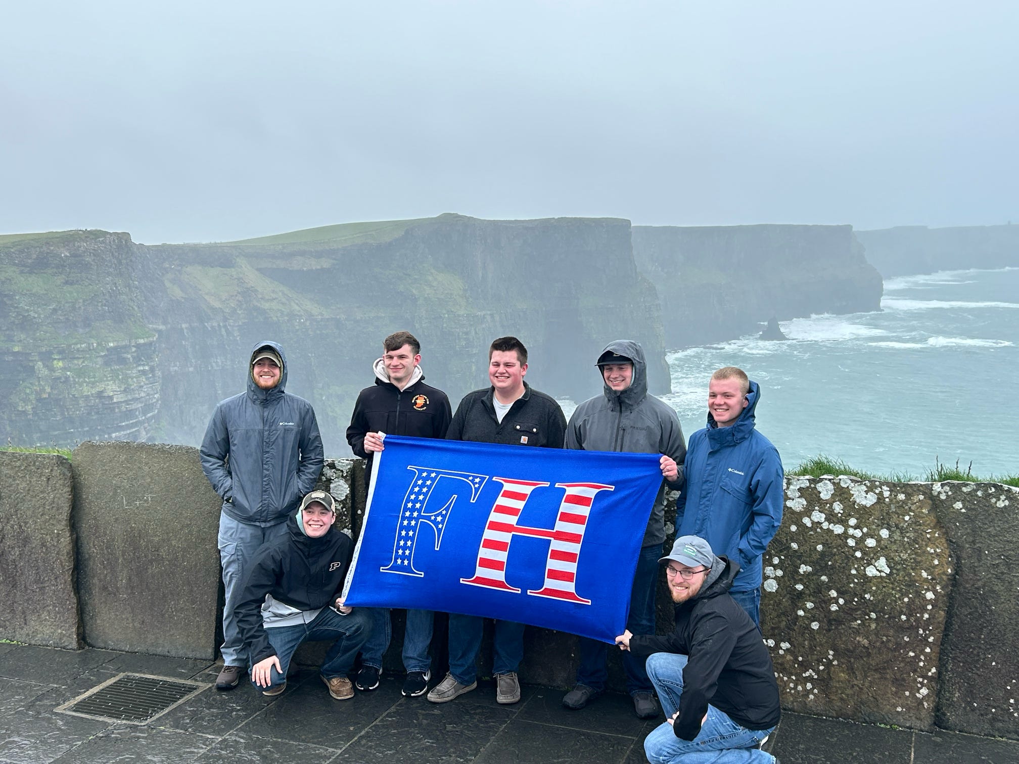 Men in rain jackets holding a FarmHouse fraternity flag with an American flag motif at coastal cliffs by the rough sea.