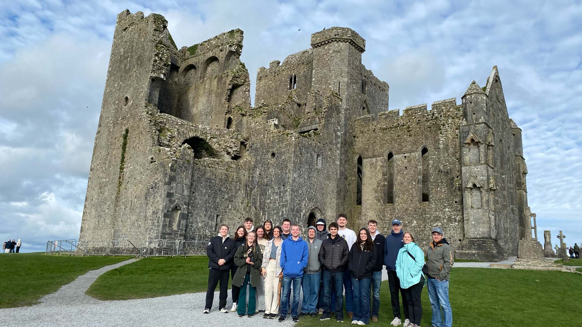 Study-abroad group posing together on the grass in front of a large medieval stone castle ruin under a cloudy sky.