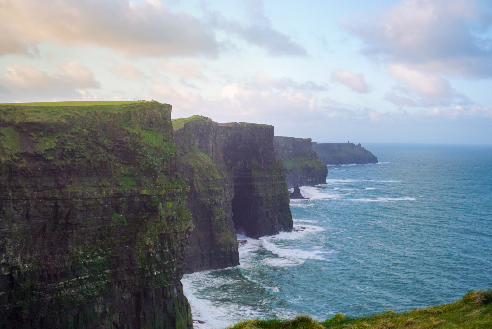 Dramatic sea cliffs with grassy tops overlooking churning blue ocean waves under a cloudy sky.