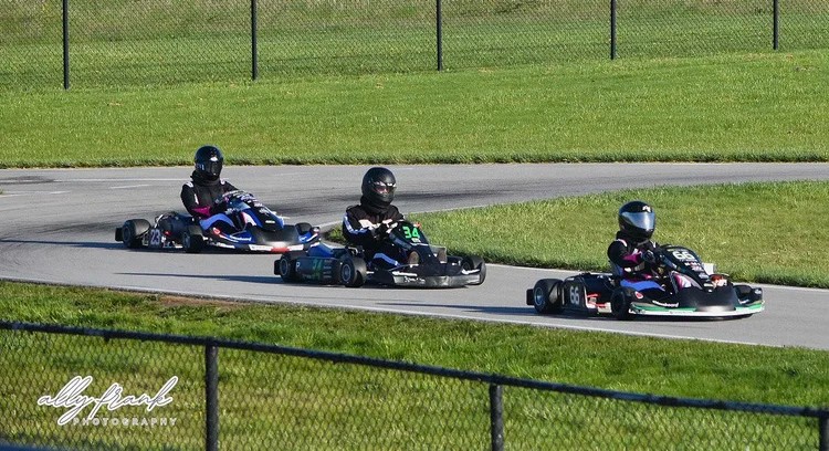 Three helmeted go-kart drivers race along a paved track with grass infield and a chain-link fence.