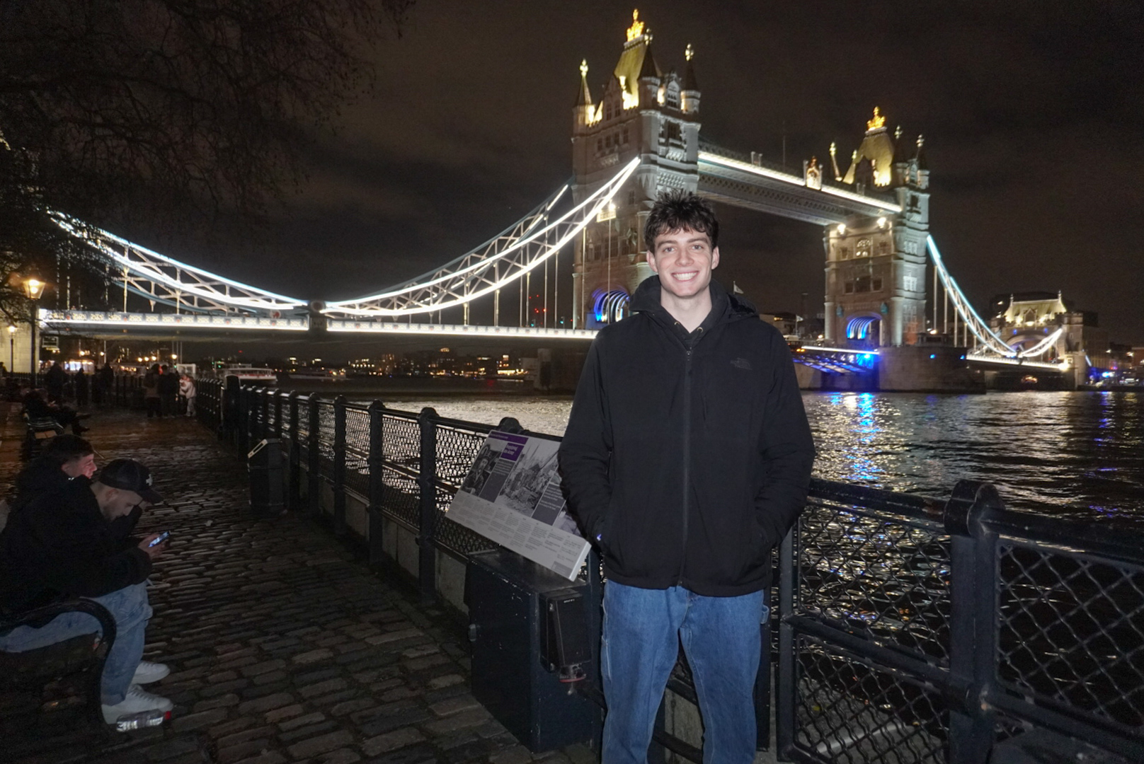 Owen in a black jacket by the River Thames with the illuminated Tower Bridge in the background at night.
