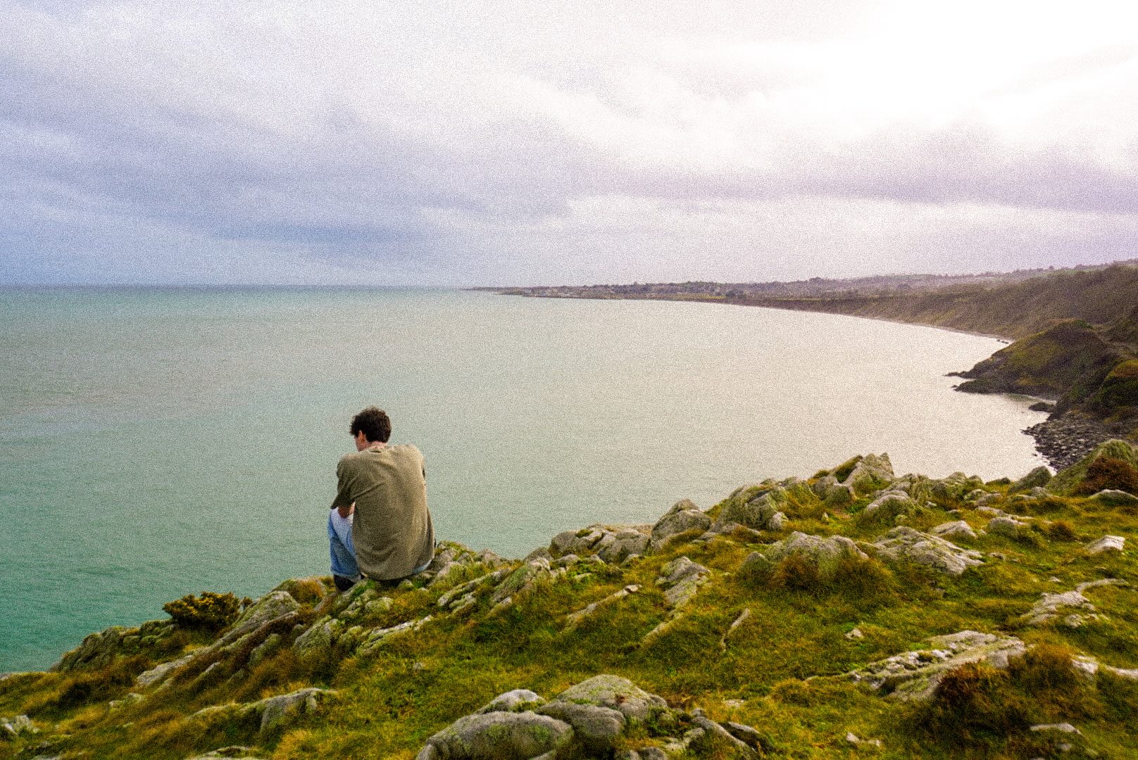 Owen sits on grassy rocky cliffs overlooking a calm sea and curving coastline under a cloudy sky.