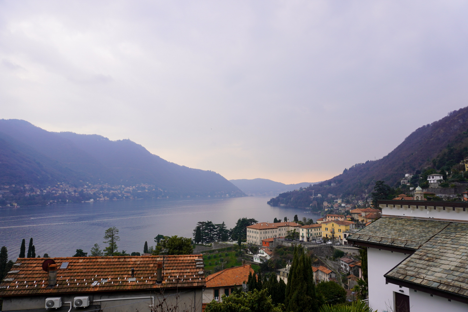 Lakeside village with terracotta roofs and pastel buildings beside a calm lake, misty mountains beyond.