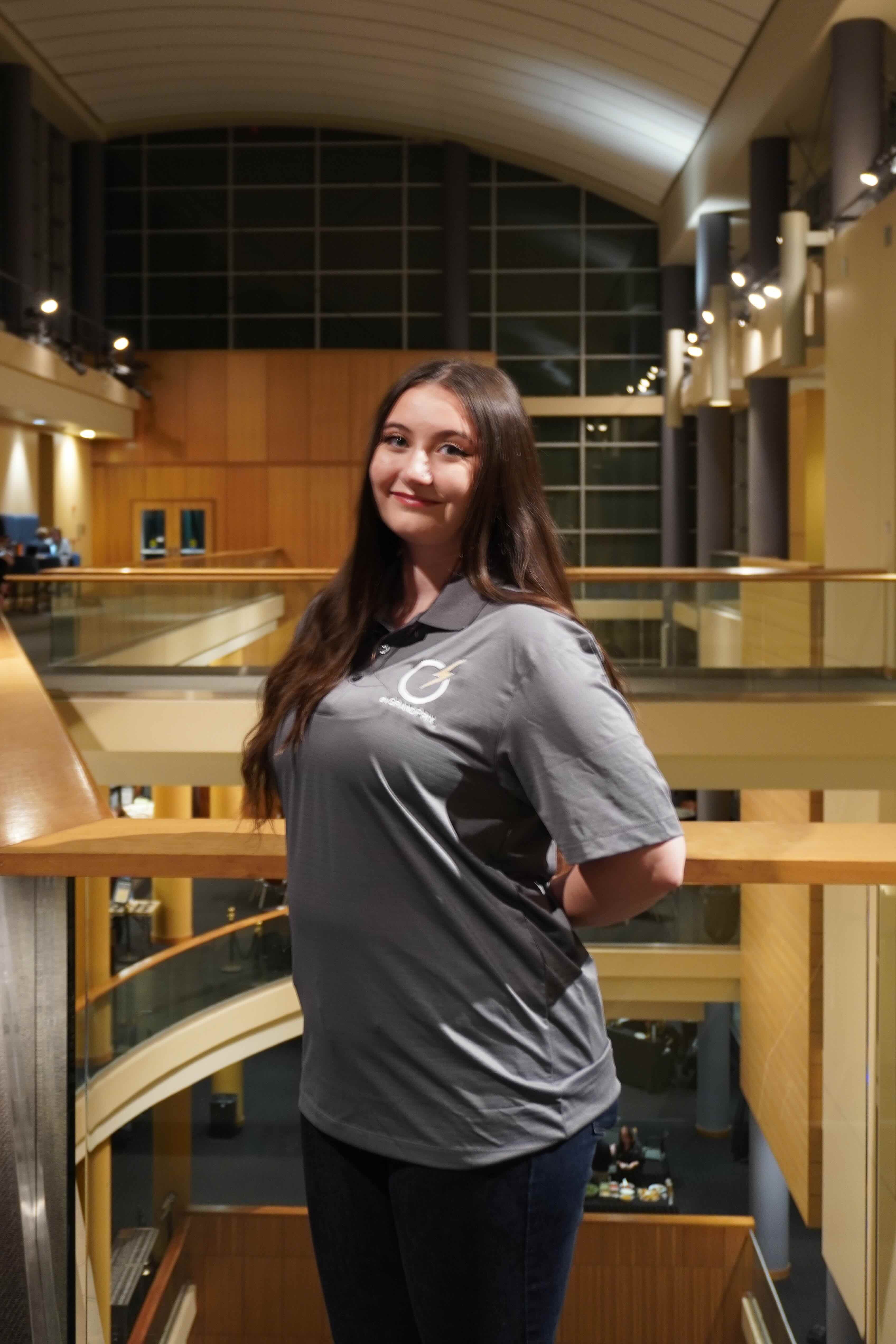 Emma is in a gray polo, smiling at the camera while standing by a railing inside a multi-level atrium.