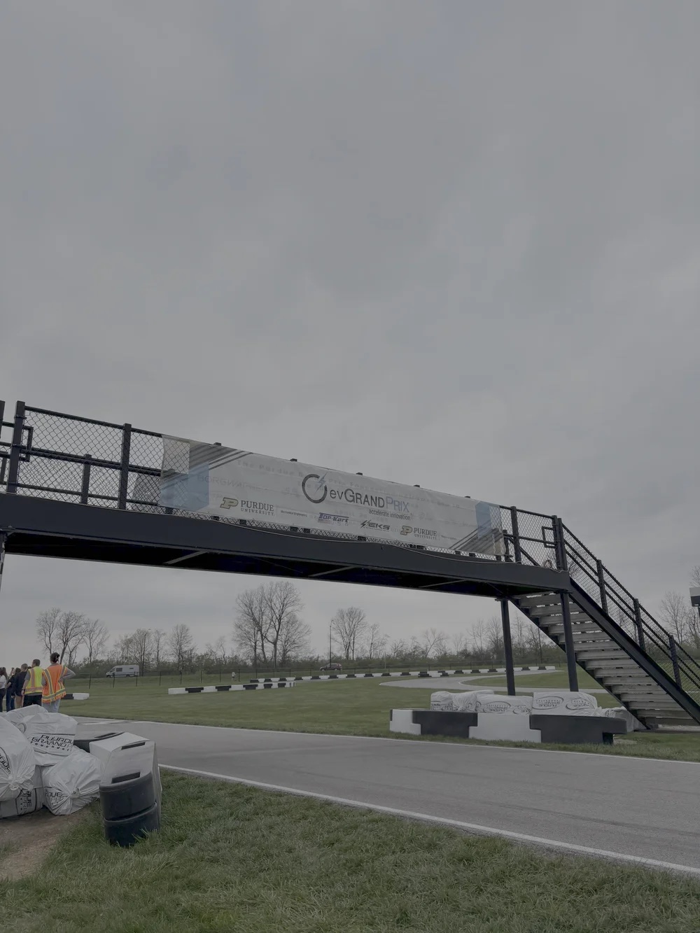 Elevated pedestrian bridge with banner over a go-kart track, stairs to the right and a few people standing by the track.