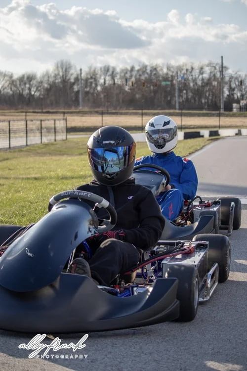 Two go-kart drivers on a paved track: lead driver in a black kart with reflective helmet, second in blue with white helmet.