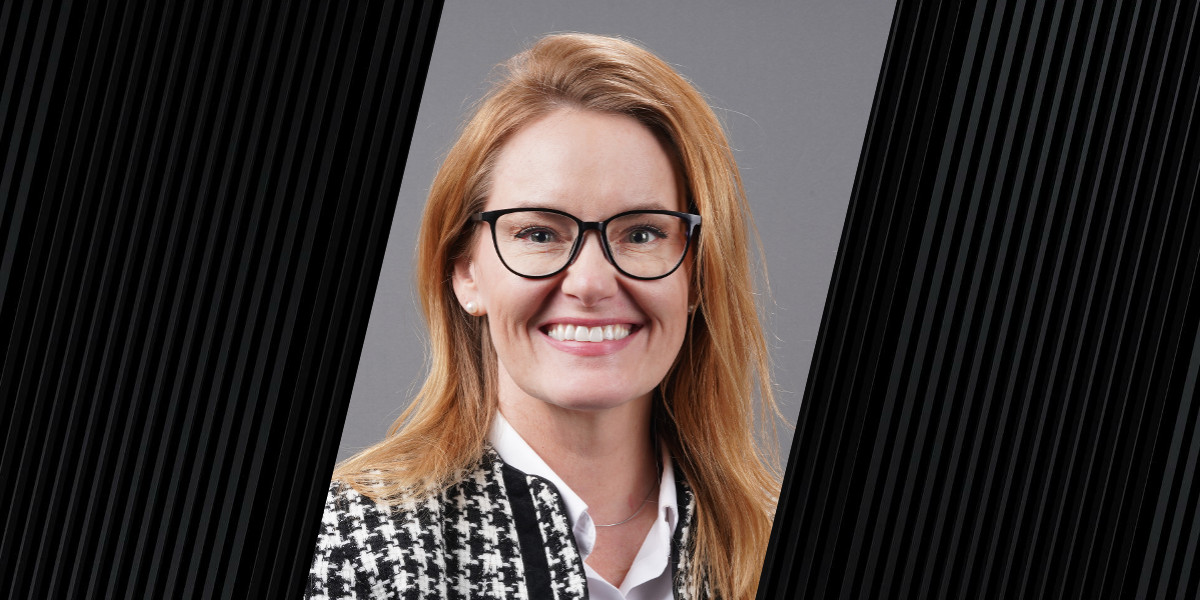 Professional headshot of a woman with shoulder-length light brown hair, wearing black-rimmed glasses, a white blouse, and a black-and-white houndstooth jacket. She is smiling in front of a gray background with black diagonal graphic borders on each side.