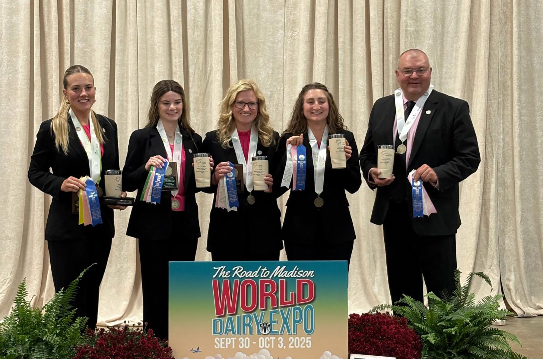 Four young women and one man, all dressed in black suits with pink shirts, stand together smiling and holding trophies, medals, and ribbons. They are posed behind a sign that reads “The Road to Madison, World Dairy Expo, Sept 30–Oct 3, 2025.” The group appears to have competed successfully, with multiple blue ribbons and awards displayed.