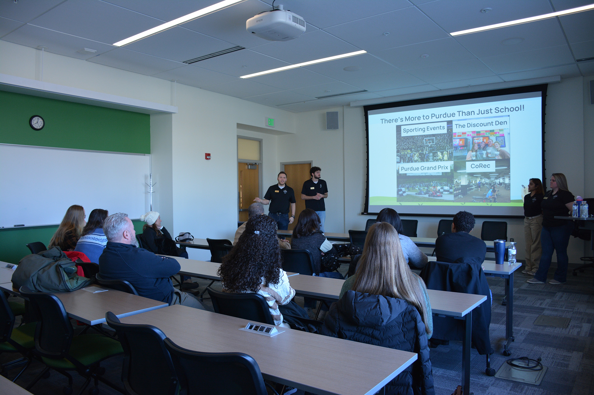 A group of prospective students and guests sit in a classroom while presenters stand at the front giving a Purdue University information session, with a slide on the screen highlighting campus activities and student life opportunities.