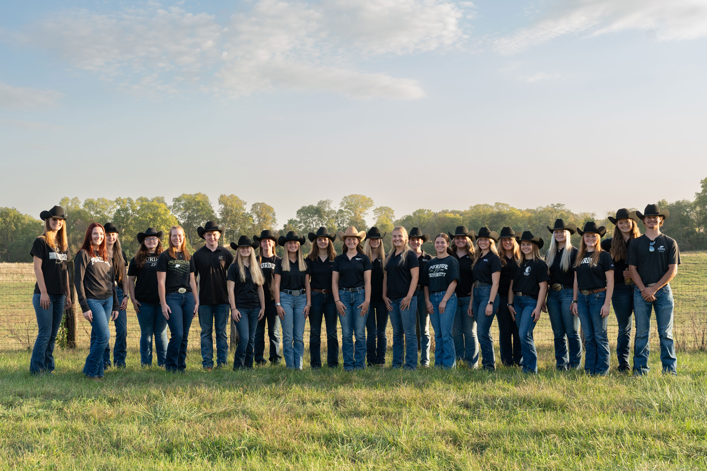 The Purdue Western Equestrian Team stands in a row outdoors in front of a fence line, wearing matching jeans, black shirts, and cowboy hats. Trees and open pasture fill the background under a partly cloudy sky.