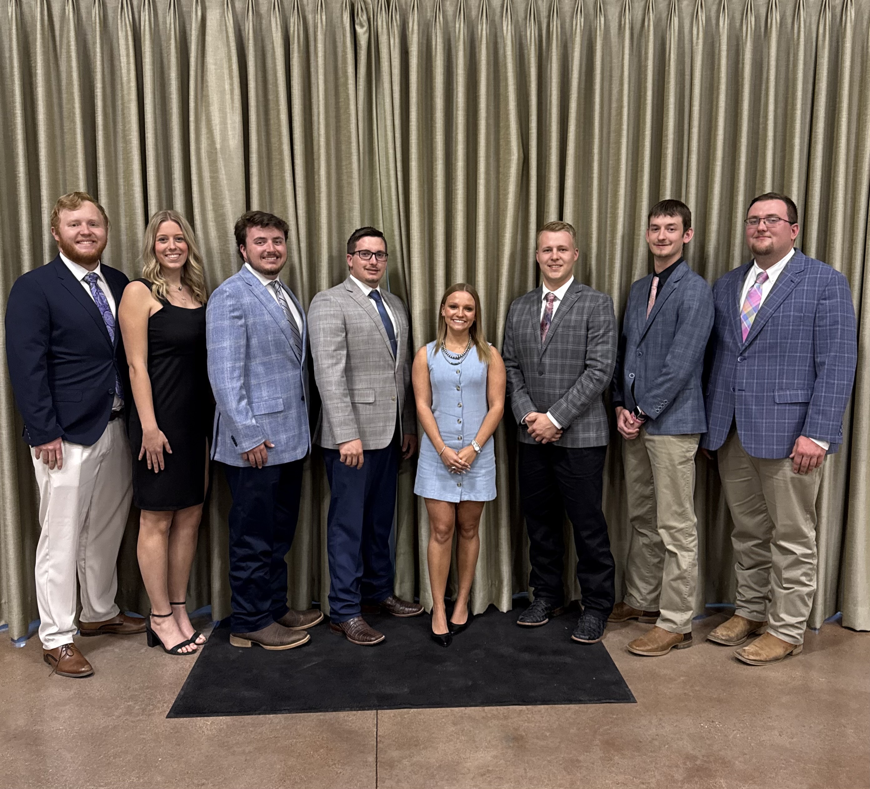 The image shows a group of eight people standing in front of a curtain. The individuals are dressed formally, with the men wearing suits and ties, and the women wearing dresses.