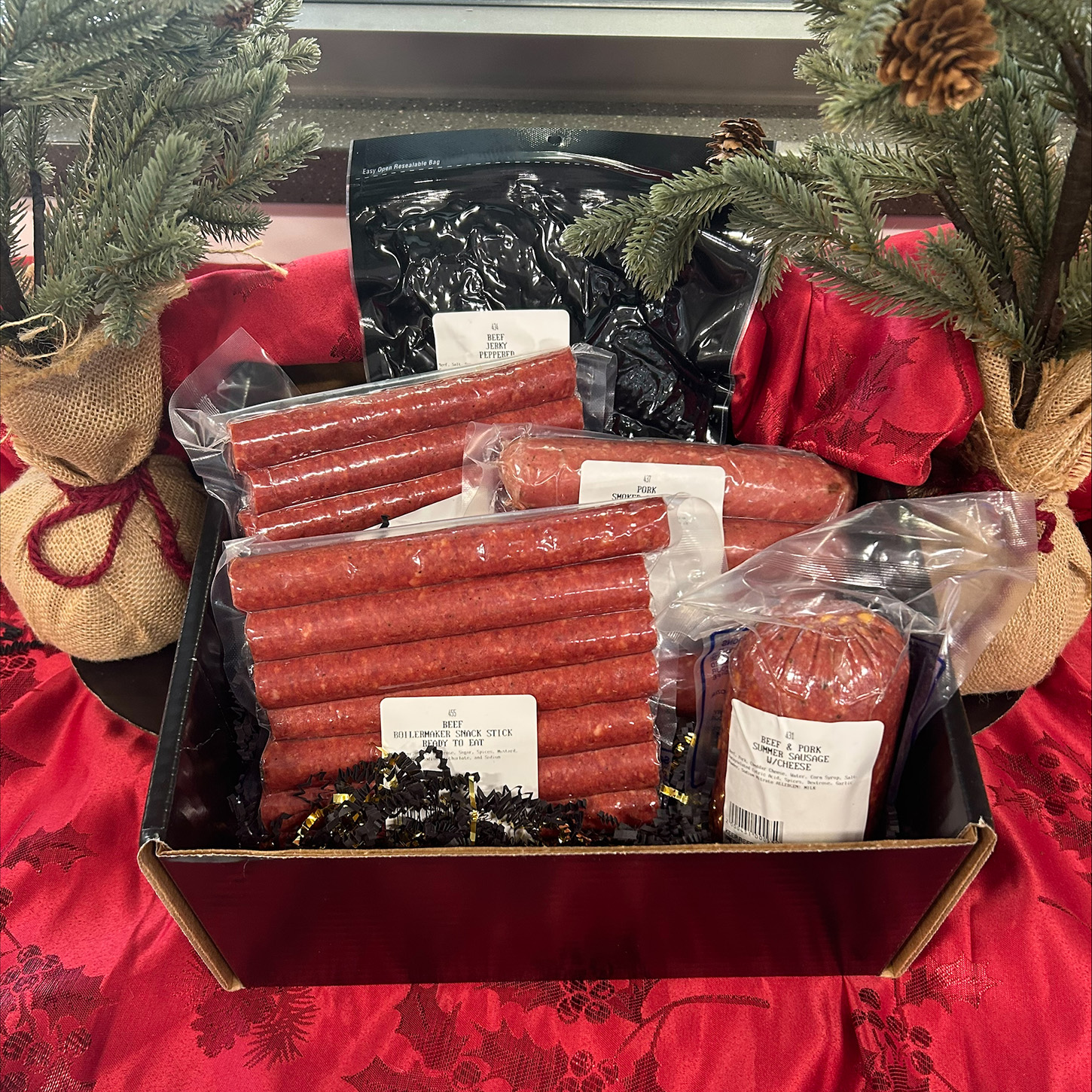 A holiday-themed gift box filled with assorted vacuum-sealed meat products, including beef and pork summer sausage and multiple packs of beef snack sticks. The items are arranged neatly in black crinkle paper inside a red box. The display is set on a red tablecloth and framed by two small burlap-wrapped evergreen decorations with pinecones.