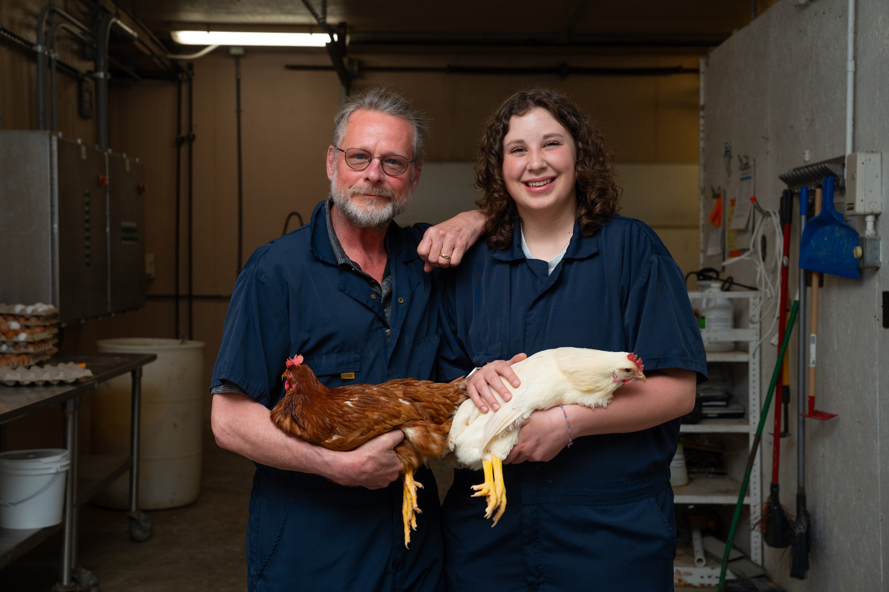 Two people wearing navy blue coveralls stand side by side in a poultry facility, each holding a chicken. The man on the left is holding a brown hen, and the woman on the right is holding a white hen. Both are smiling at the camera, with equipment and supplies visible in the background.
