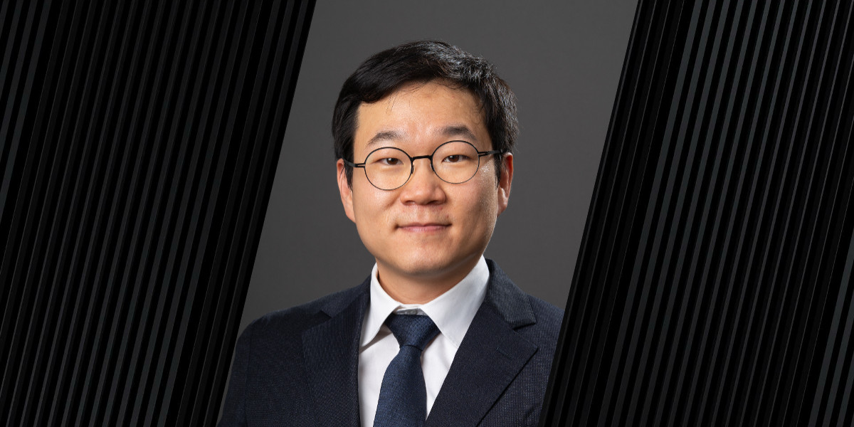 A person wearing a dark suit, white shirt, and dark tie stands against a plain dark background with vertical black stripes on both sides.