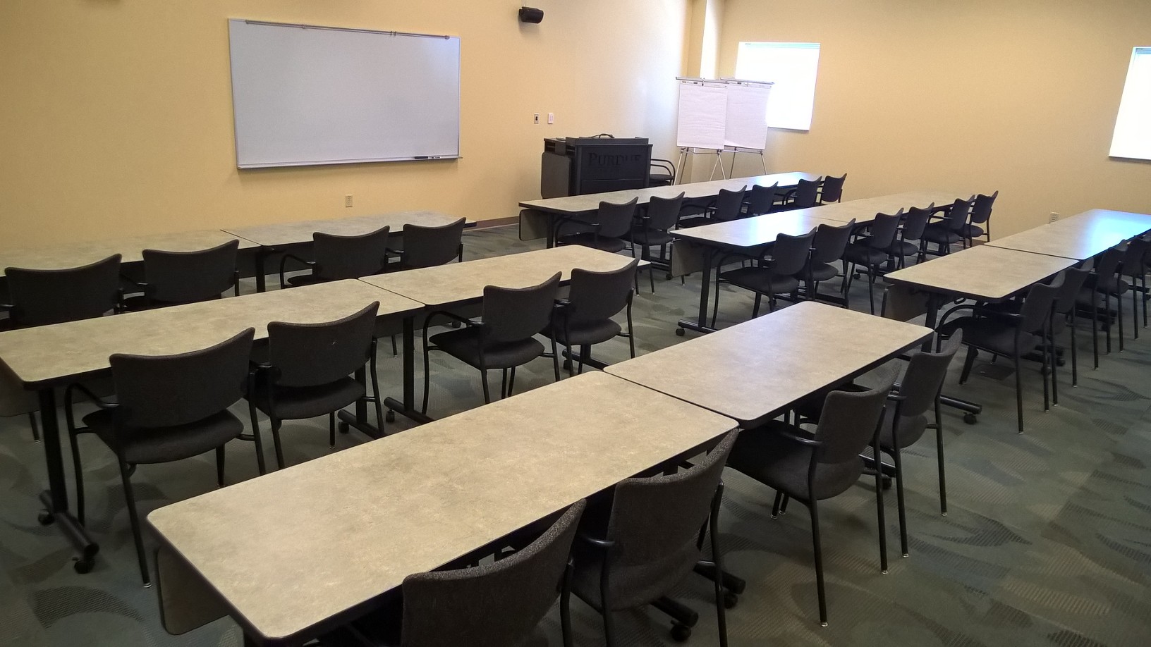 Room 117 set up in classroom style seating facing the front. Room can sit up to 32 guests in fixed, cushioned chairs. Seating faces towards the front white board and AV podium. There is a projector screen that can be rolled up or down for presentations.