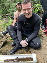 Zach Kellogg sitting on ground by river with live hellbender in water tube.