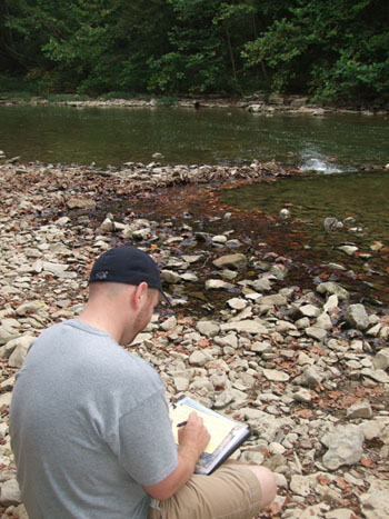 Staff member sitting on the blue river bank taking notes.